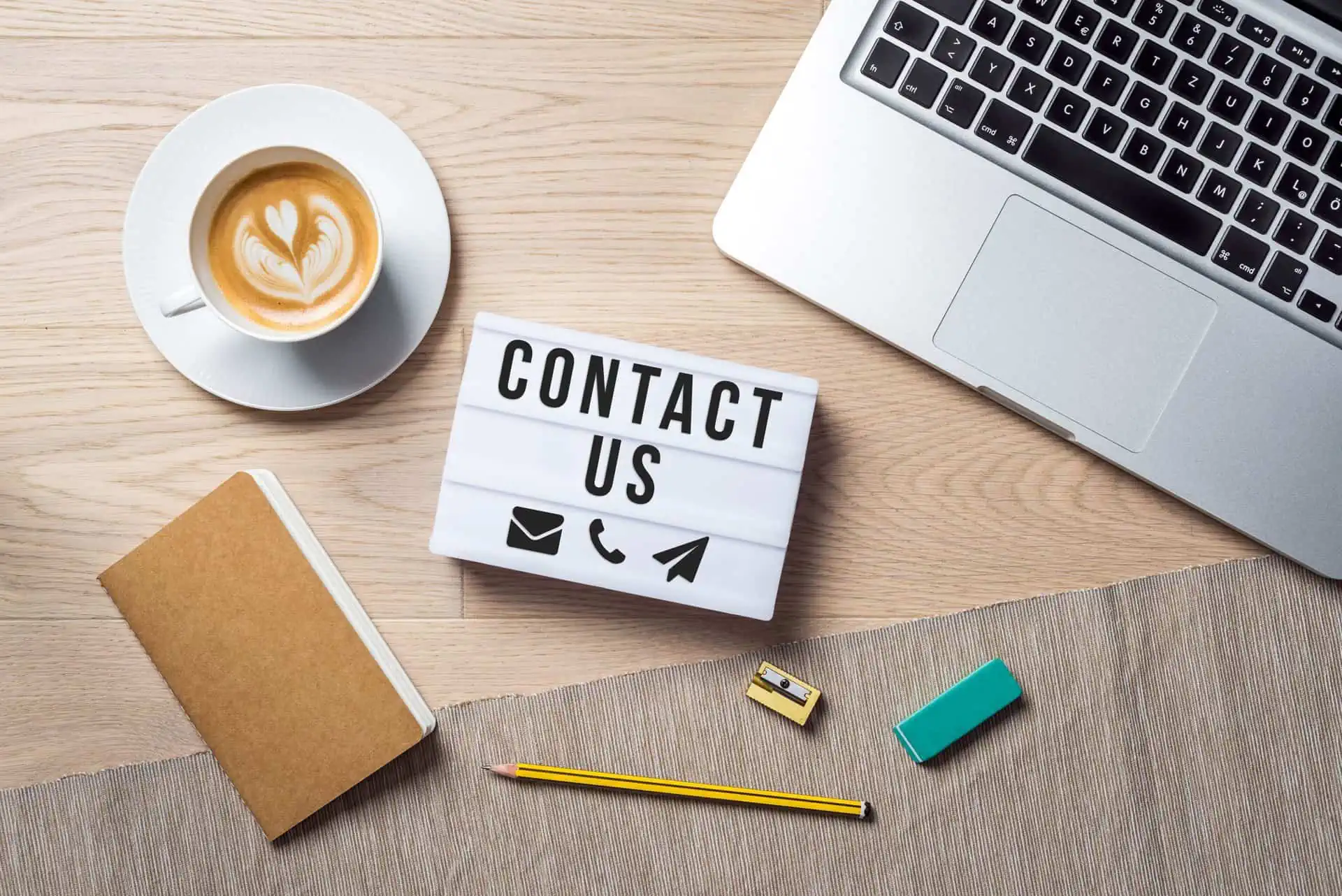 Contact us writing with emoji in lightbox lying on wooden desk with office supply and coffee cup as flat lay from bird's eye view